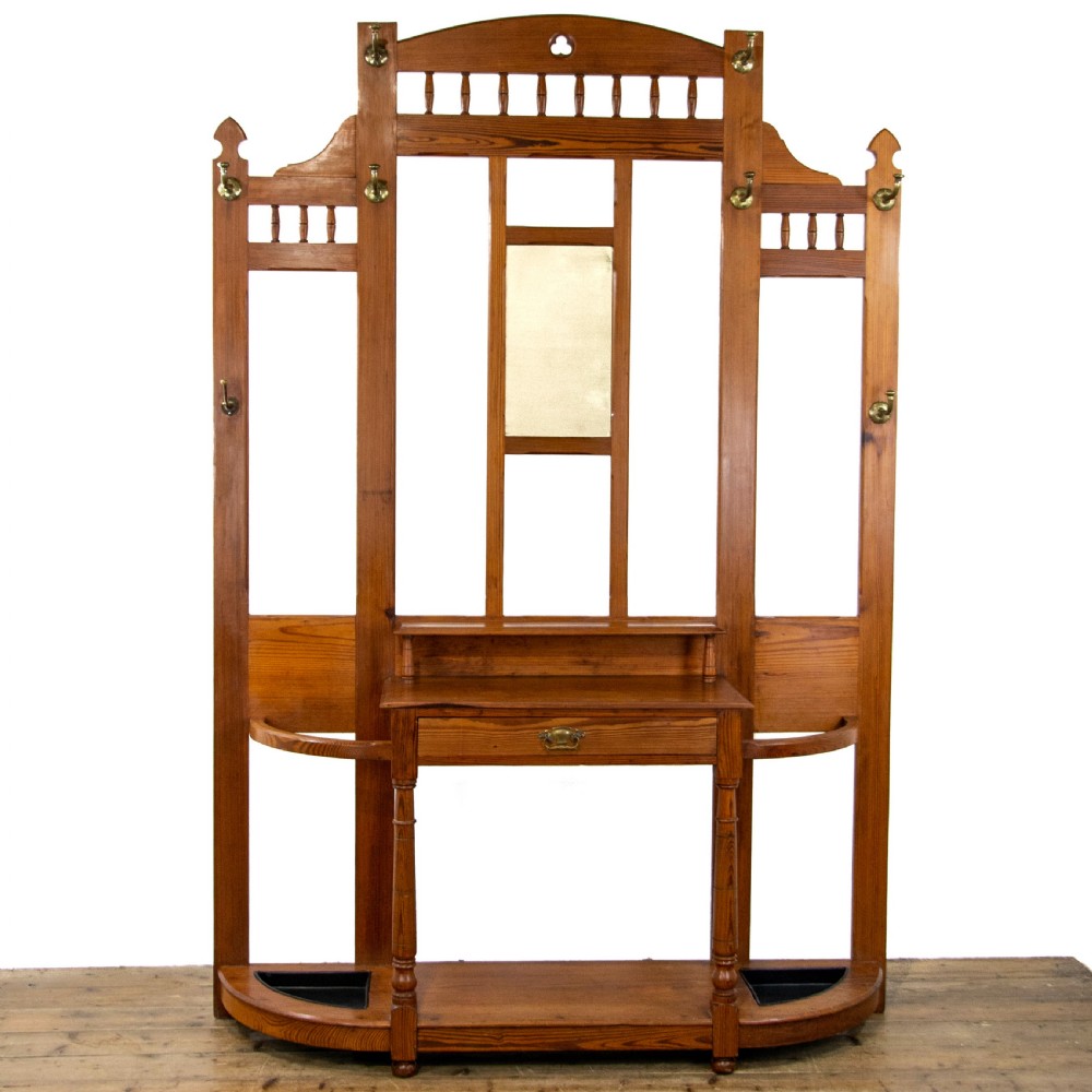 A tall Victorian pitch pine hall stand featuring a central mirror, brass coat hooks, a small drawer unit, and integrated umbrella racks with drip trays.