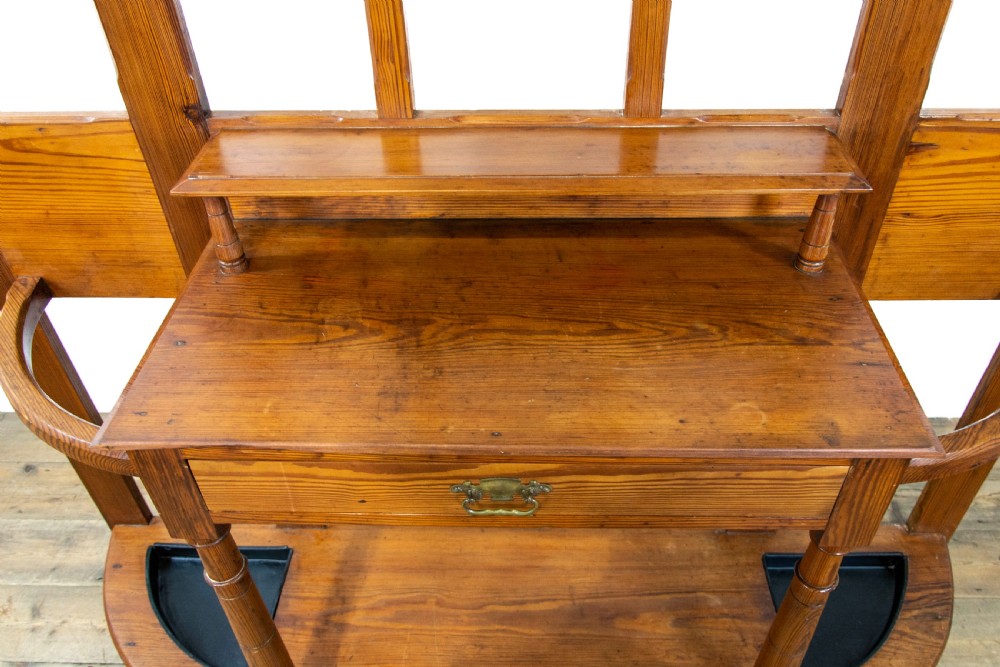 A tall Victorian pitch pine hall stand featuring a central mirror, brass coat hooks, a small drawer unit, and integrated umbrella racks with drip trays. View VII