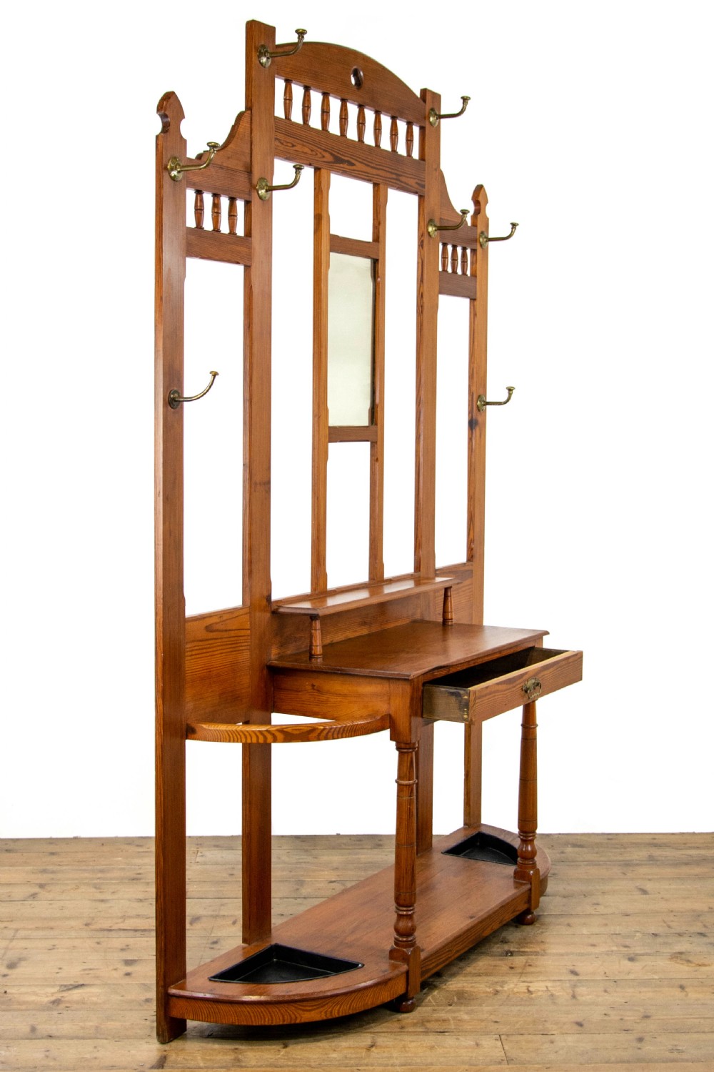 A tall Victorian pitch pine hall stand featuring a central mirror, brass coat hooks, a small drawer unit, and integrated umbrella racks with drip trays. View X