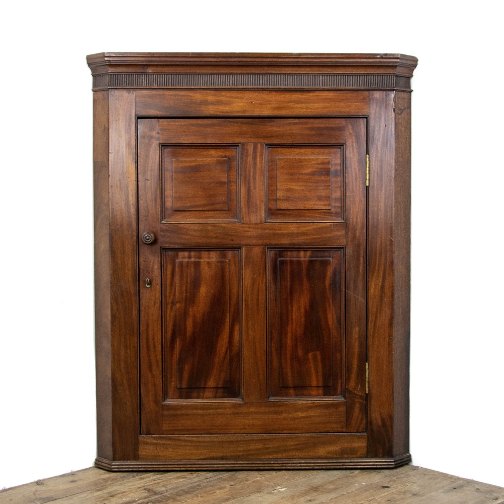 An antique mahogany corner cupboard featuring a single door with four raised flame mahogany panels, a fluted cornice, and brass hardware.