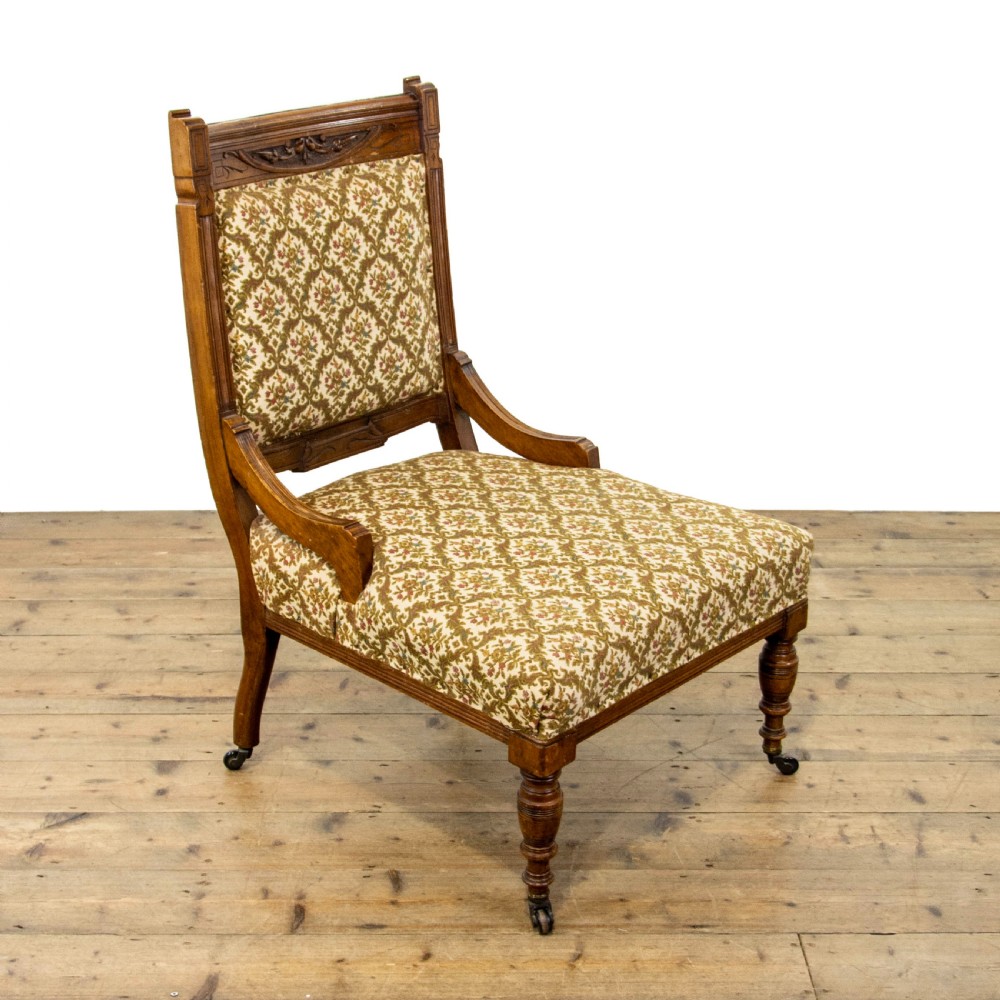An antique Edwardian low-slung chair featuring a carved timber frame, patterned textile upholstery, turned front legs, and castors.