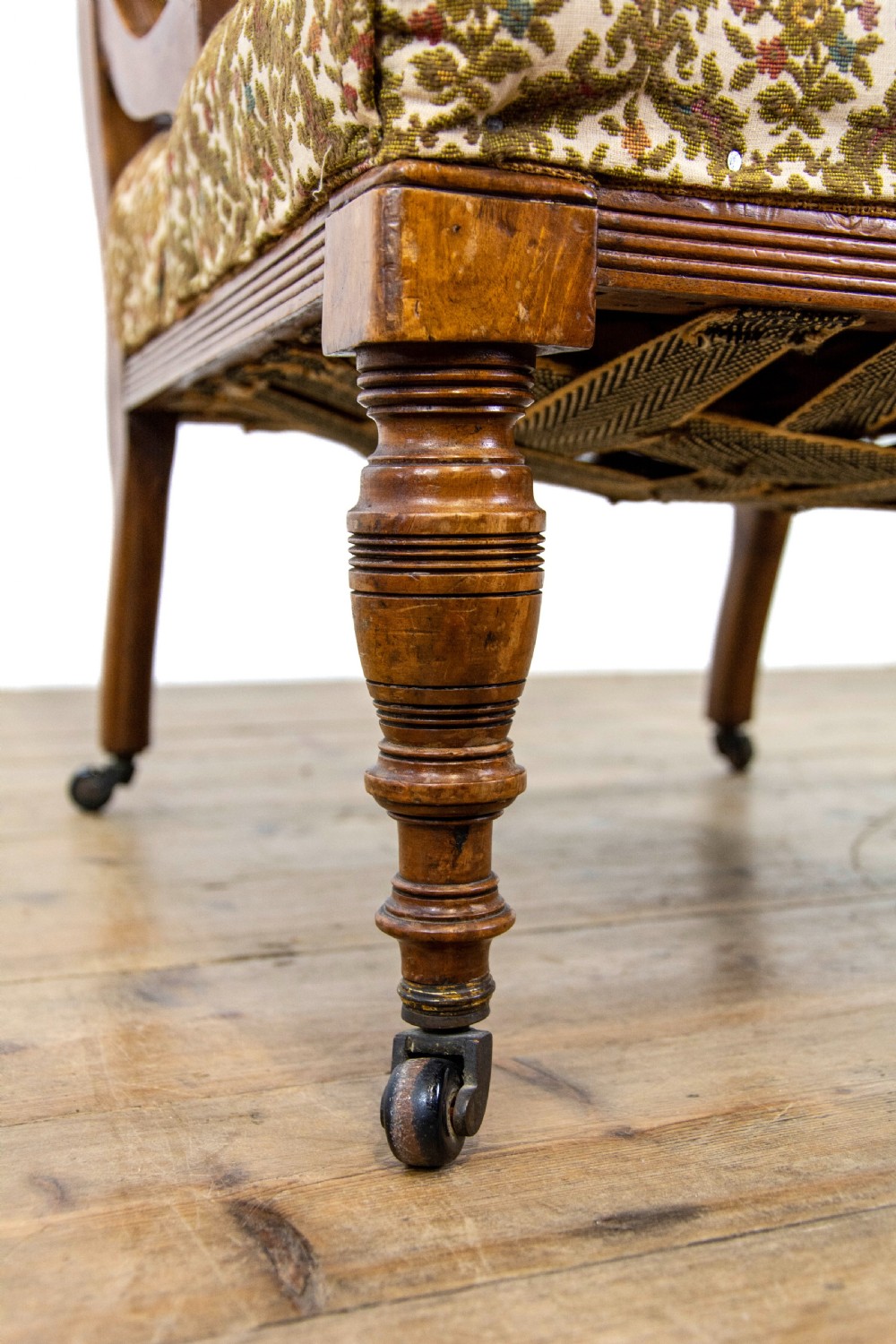 An antique Edwardian low-slung chair featuring a carved timber frame, patterned textile upholstery, turned front legs, and castors. View V