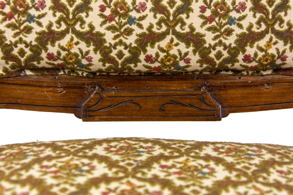 An antique Edwardian low-slung chair featuring a carved timber frame, patterned textile upholstery, turned front legs, and castors. View VI