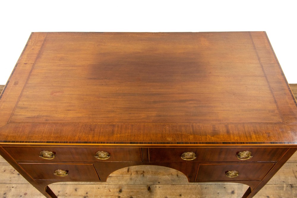 An antique Sheraton Revival mahogany side table featuring four drawers, string inlay detailing, brass ring handles, and an arched central kneehole on tapered square legs. View V
