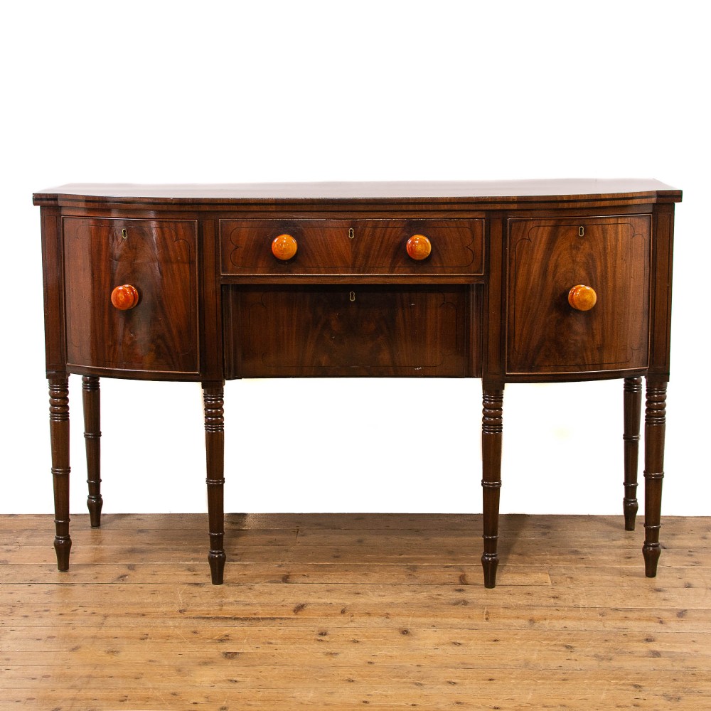 An antique mahogany bow-front sideboard featuring turned reeded legs, central drawers, and flanking cupboard doors with turned wooden knobs.