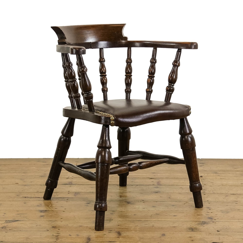 A dark wood antique smokers bow armchair featuring turned spindles, a curved crest rail, and a brown leather seat with brass studs.