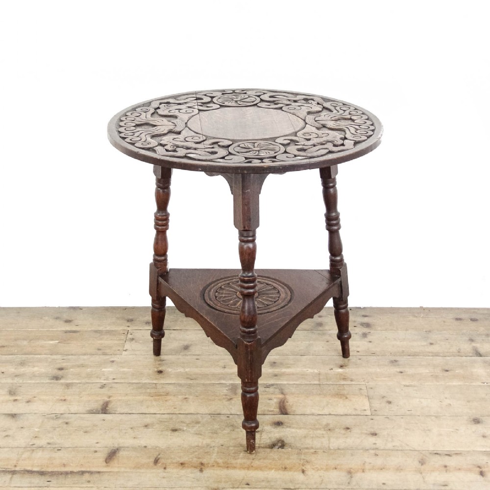 An early twentieth-century dark oak cricket table featuring a circular carved top, three turned legs, and a triangular lower shelf.