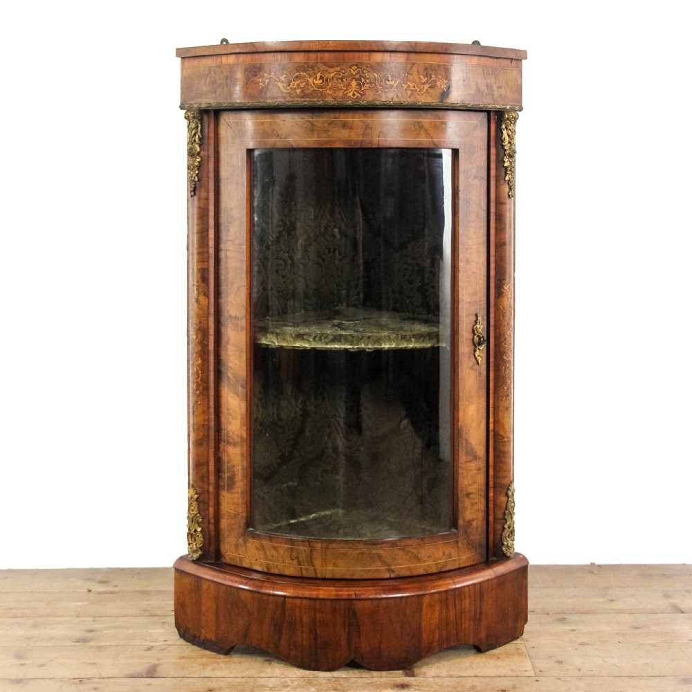 A Victorian bow front corner cupboard made of polished walnut wood, featuring floral marquetry inlay on the top frieze, gilt bronze corner mounts, and a glass-fronted door revealing a fabric-lined interior.