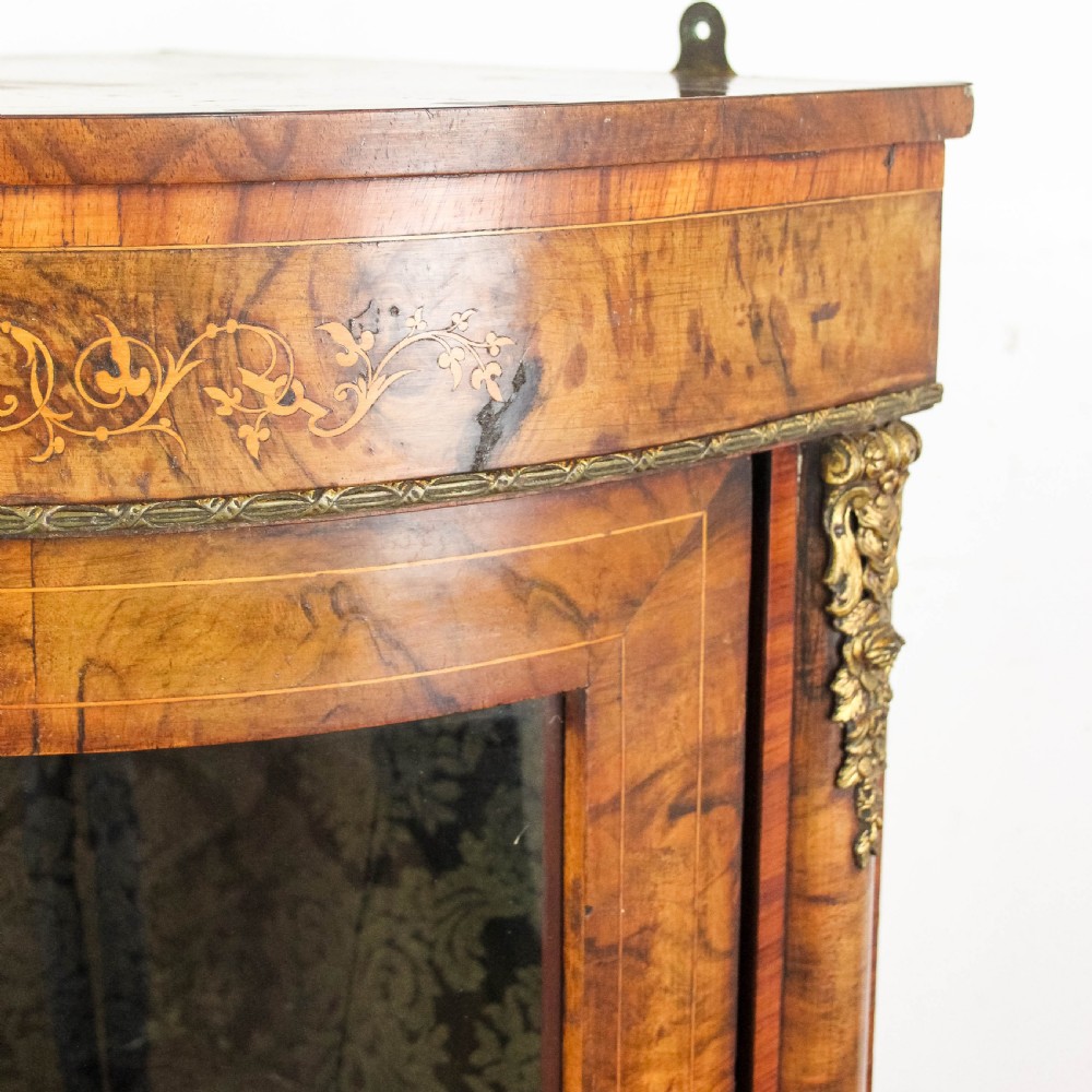 A Victorian bow front corner cupboard made of polished walnut wood, featuring floral marquetry inlay on the top frieze, gilt bronze corner mounts, and a glass-fronted door revealing a fabric-lined interior. View II