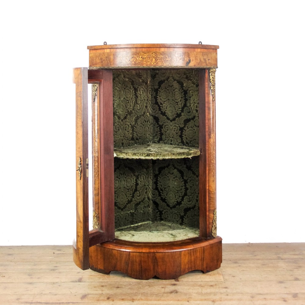 A Victorian bow front corner cupboard made of polished walnut wood, featuring floral marquetry inlay on the top frieze, gilt bronze corner mounts, and a glass-fronted door revealing a fabric-lined interior. View III