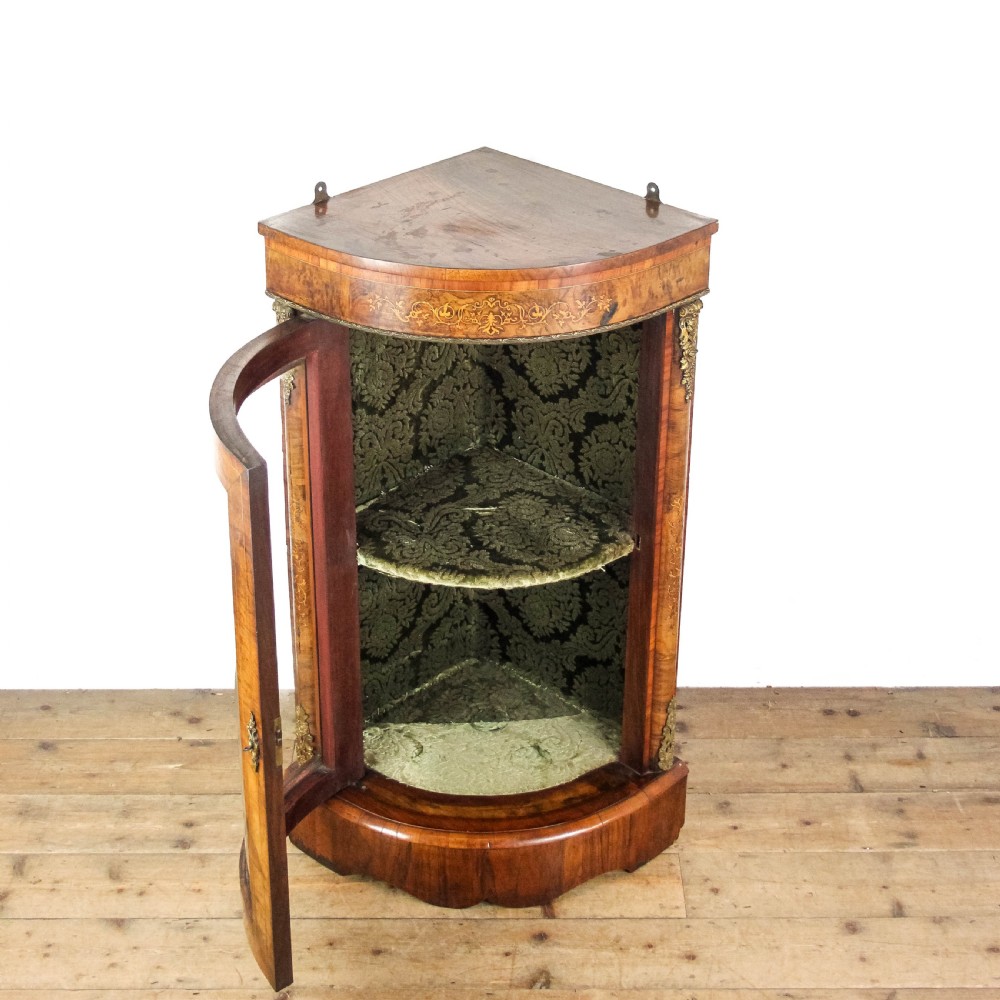 A Victorian bow front corner cupboard made of polished walnut wood, featuring floral marquetry inlay on the top frieze, gilt bronze corner mounts, and a glass-fronted door revealing a fabric-lined interior. View IV
