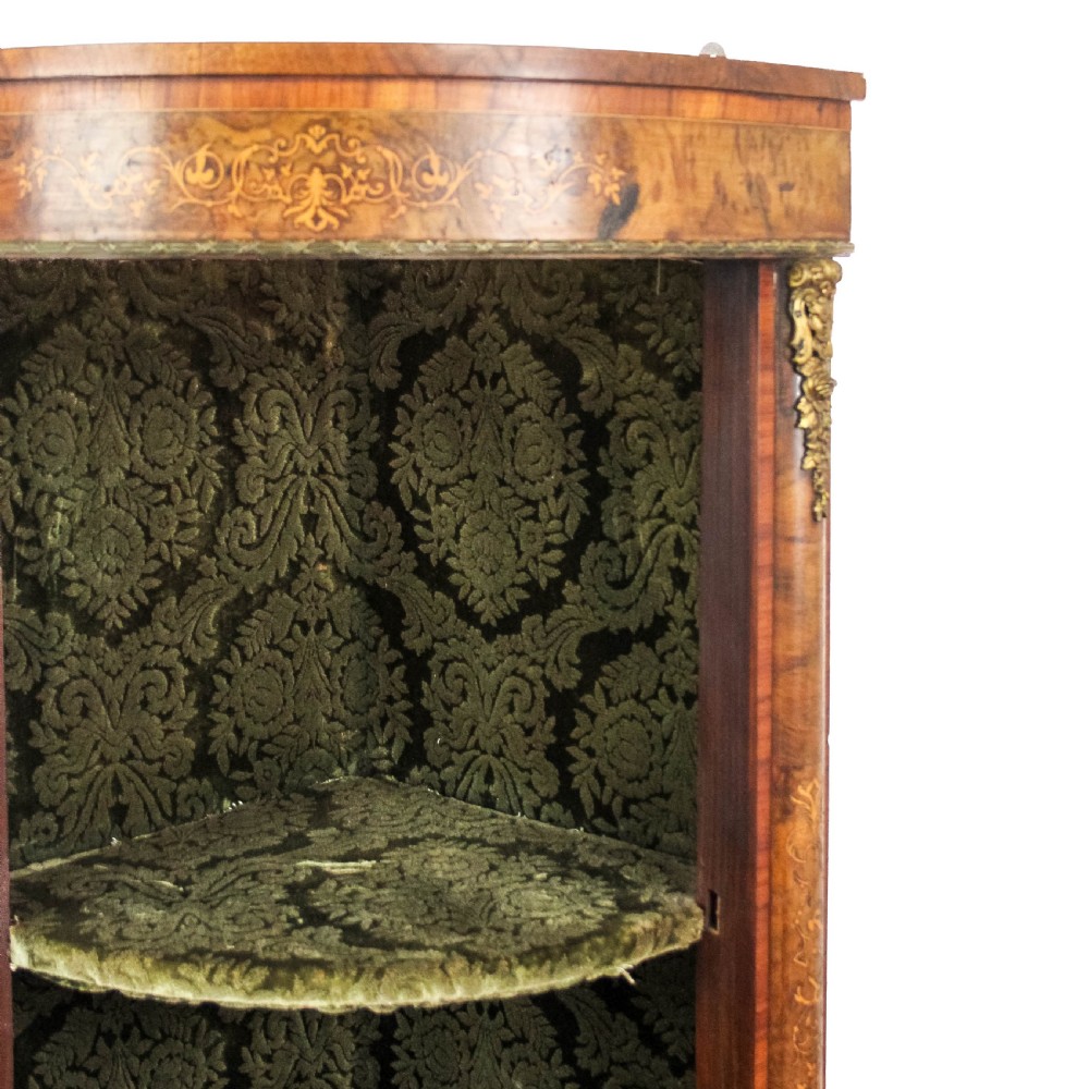A Victorian bow front corner cupboard made of polished walnut wood, featuring floral marquetry inlay on the top frieze, gilt bronze corner mounts, and a glass-fronted door revealing a fabric-lined interior. View V