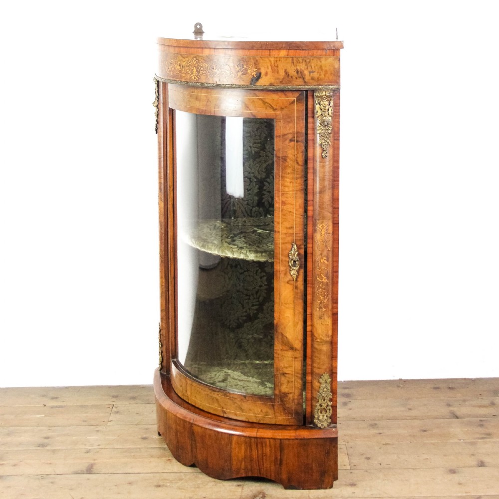 A Victorian bow front corner cupboard made of polished walnut wood, featuring floral marquetry inlay on the top frieze, gilt bronze corner mounts, and a glass-fronted door revealing a fabric-lined interior. View VI