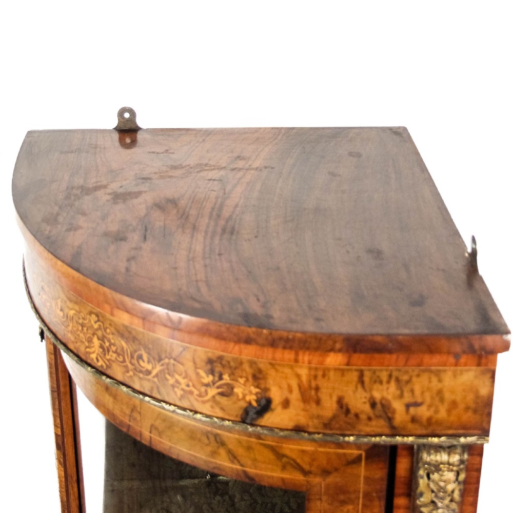 A Victorian bow front corner cupboard made of polished walnut wood, featuring floral marquetry inlay on the top frieze, gilt bronze corner mounts, and a glass-fronted door revealing a fabric-lined interior. View VII