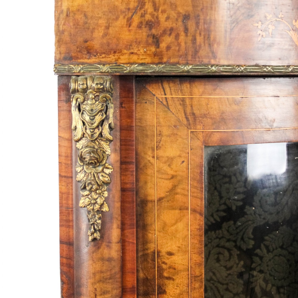 A Victorian bow front corner cupboard made of polished walnut wood, featuring floral marquetry inlay on the top frieze, gilt bronze corner mounts, and a glass-fronted door revealing a fabric-lined interior. View VIII
