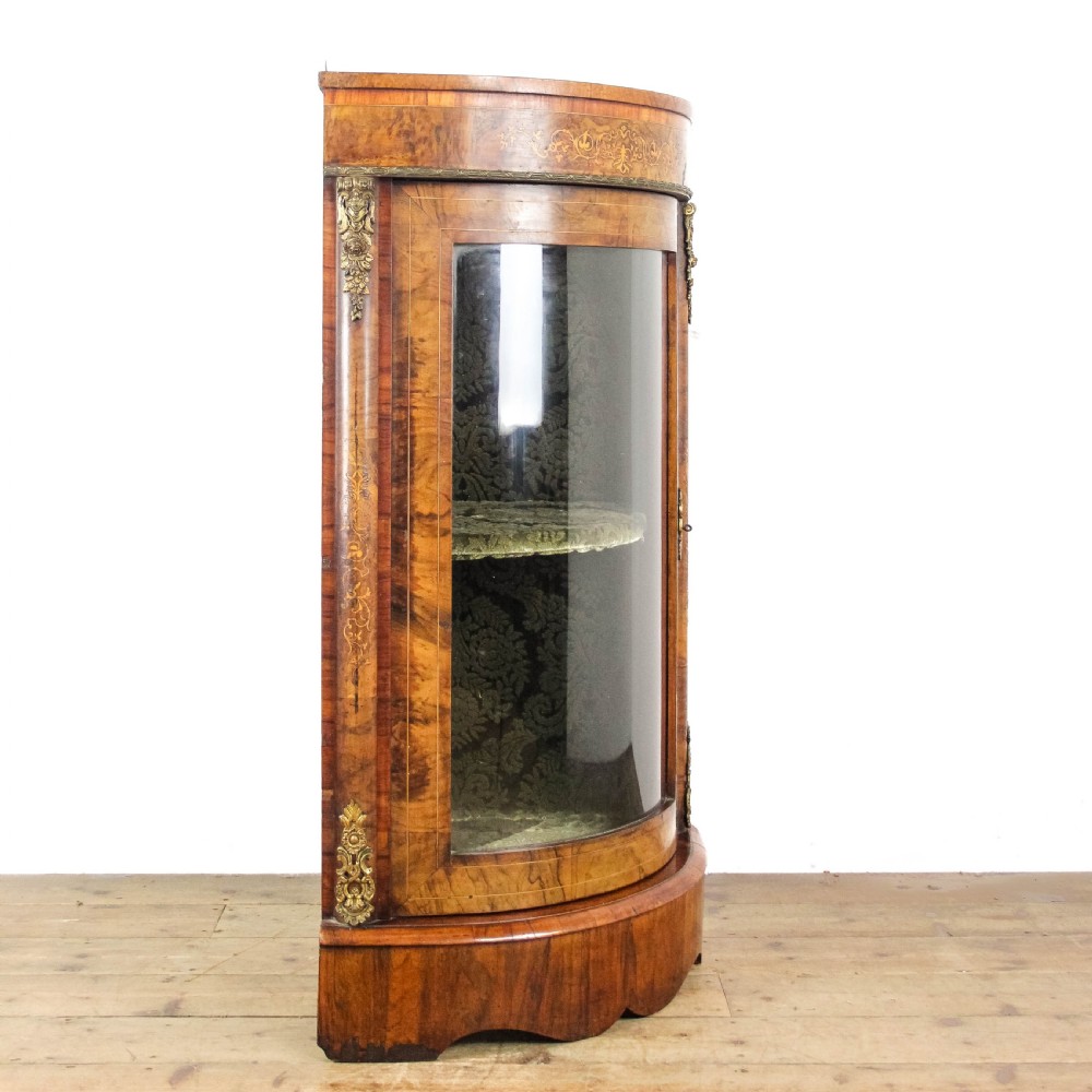 A Victorian bow front corner cupboard made of polished walnut wood, featuring floral marquetry inlay on the top frieze, gilt bronze corner mounts, and a glass-fronted door revealing a fabric-lined interior. View IX