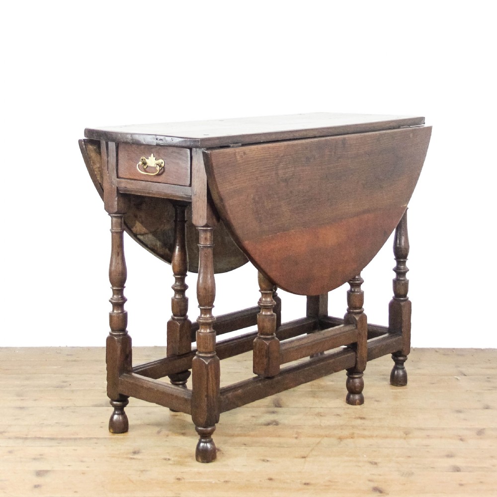 An antique dark oak gateleg table featuring two hinged drop leaves, turned legs, lower stretchers, and a single end drawer with a brass handle.