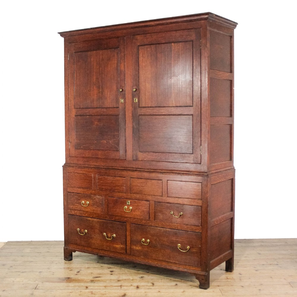 An antique dark oak linen press cabinet featuring two panelled upper doors above a lower chest containing seven drawers with brass swan-neck handles. View II