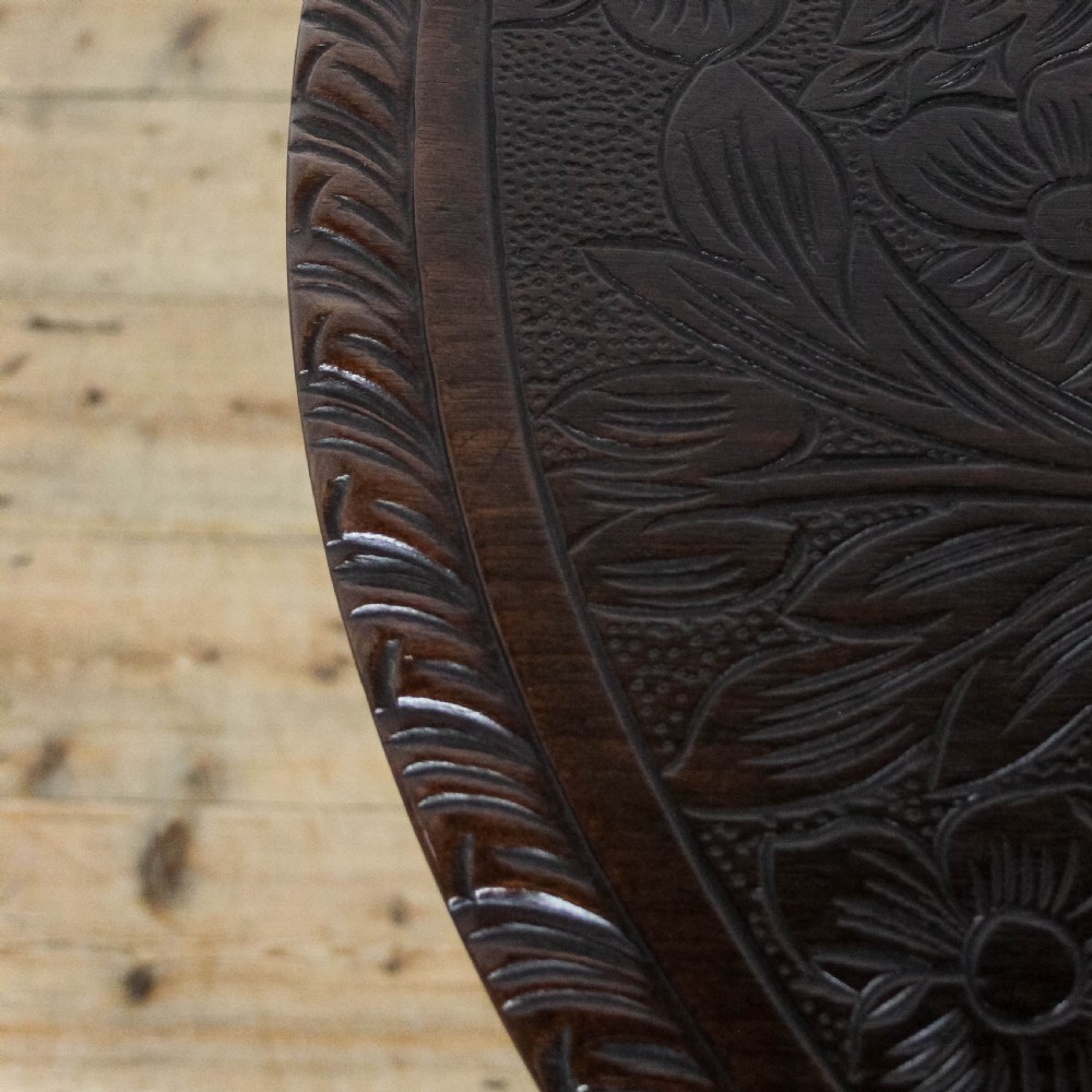 A dark mahogany tilt-top tripod table featuring an intricately carved circular top with a floral vase motif, supported on three carved legs. View V