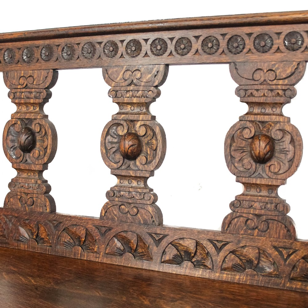 A dark carved oak hall bench featuring turned front legs, shaped armrests, and an open carved backrest. View IX