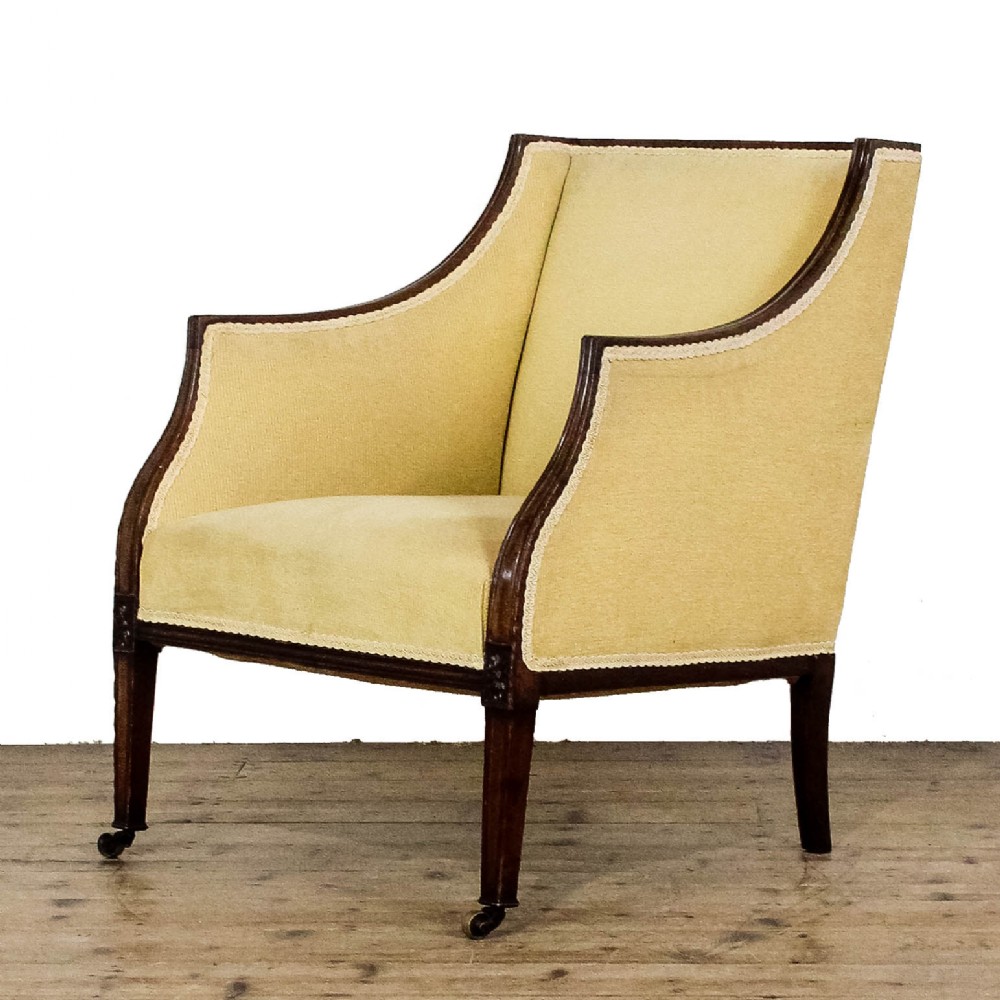 An Edwardian armchair with pale yellow fabric upholstery, a dark mahogany frame with sweeping armrests, square tapered front legs, and brass castors.