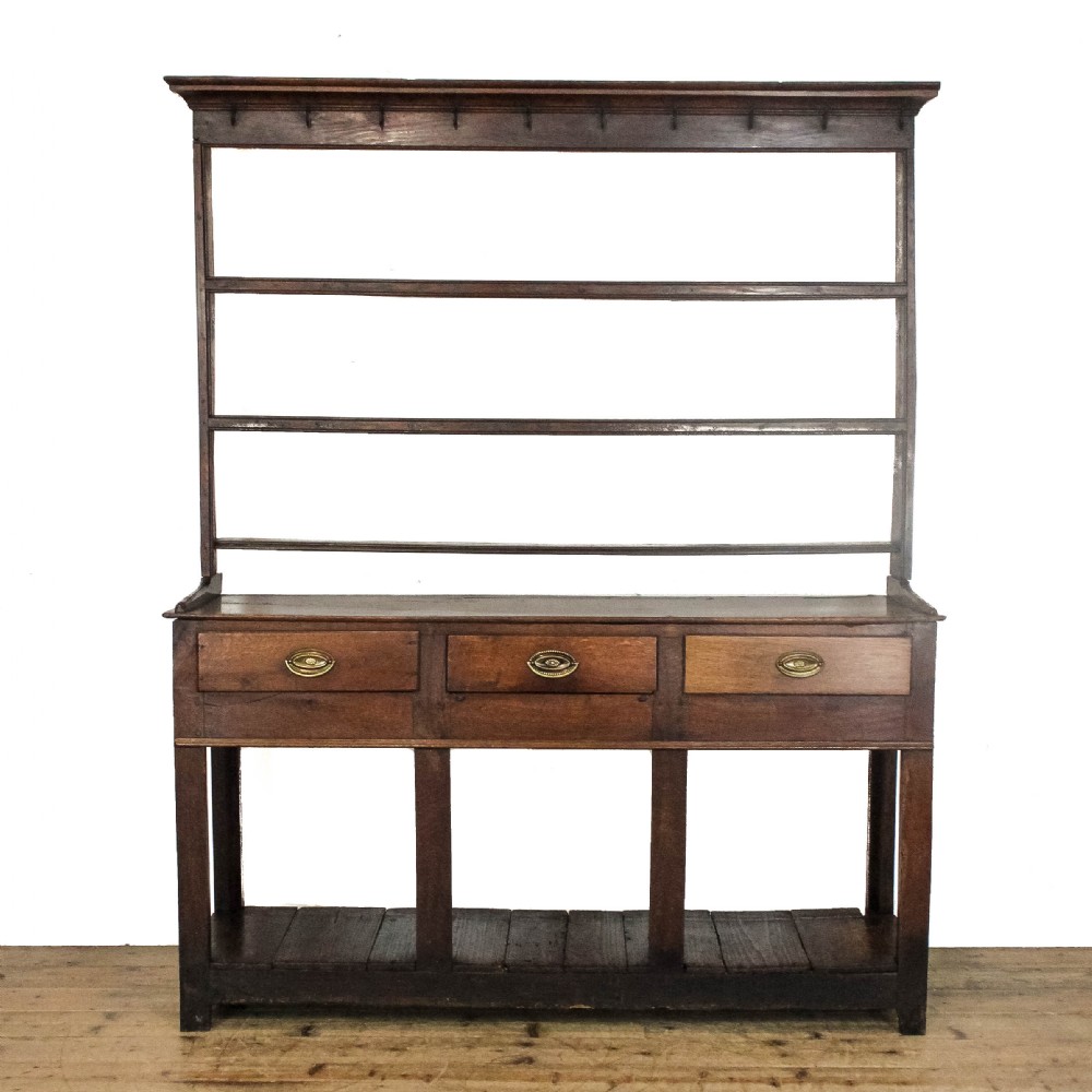 An antique dark oak open-potboard dresser featuring an upper three-tier plate rack with hooks, three drawer units with brass oval handles, and four square legs supporting a slatted lower shelf.