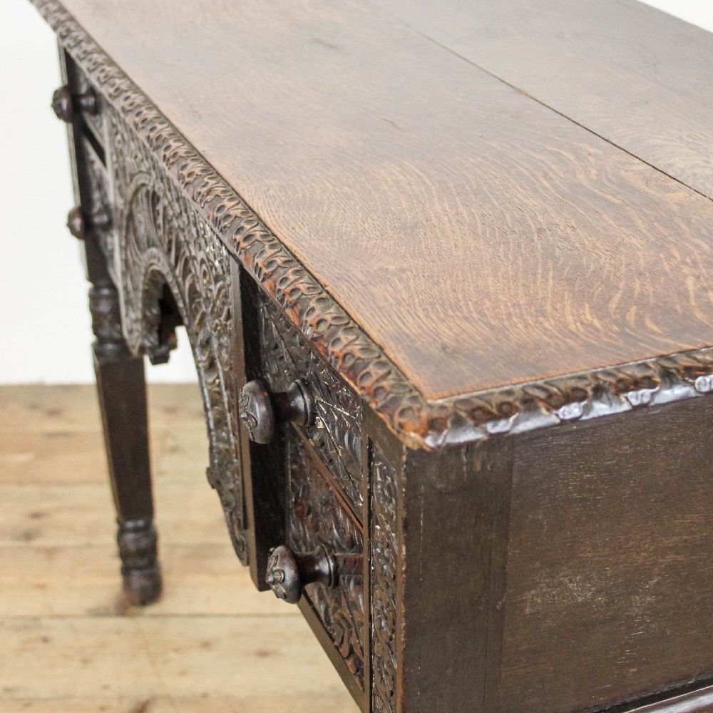 A dark-stained antique oak side table featuring intricate foliate relief carvings, a central arched kneehole, four small drawers, and turned legs. View VII