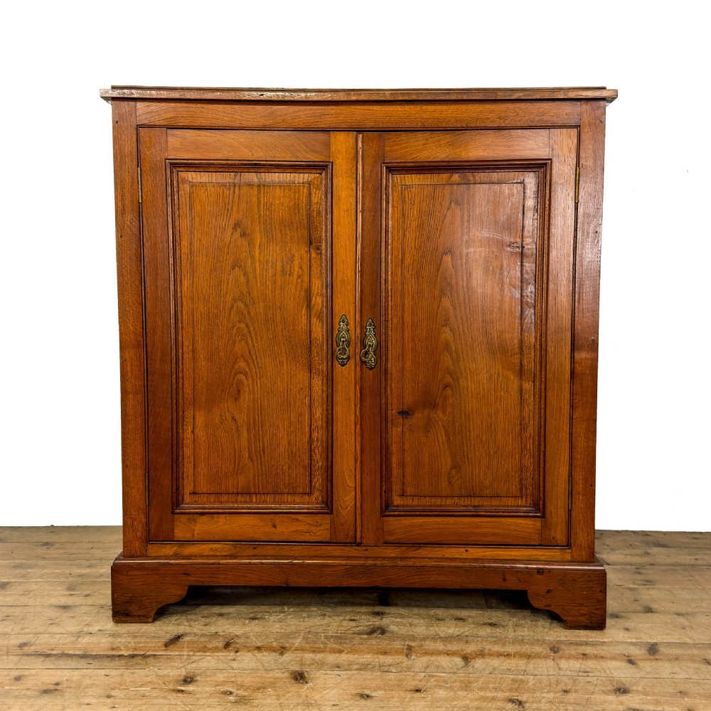 An antique warm brown oak cupboard featuring two moulded panel doors, polished brass escutcheons, and a shaped plinth base.