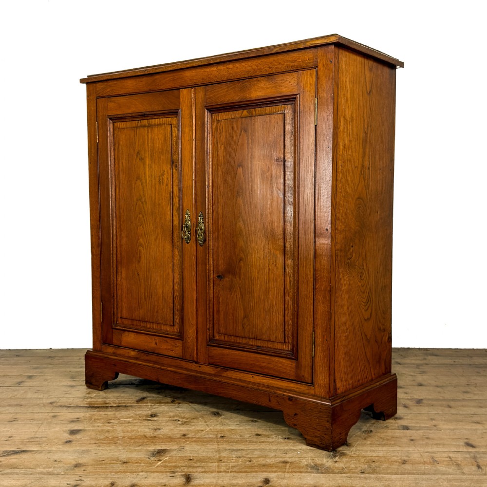 An antique warm brown oak cupboard featuring two moulded panel doors, polished brass escutcheons, and a shaped plinth base. View II
