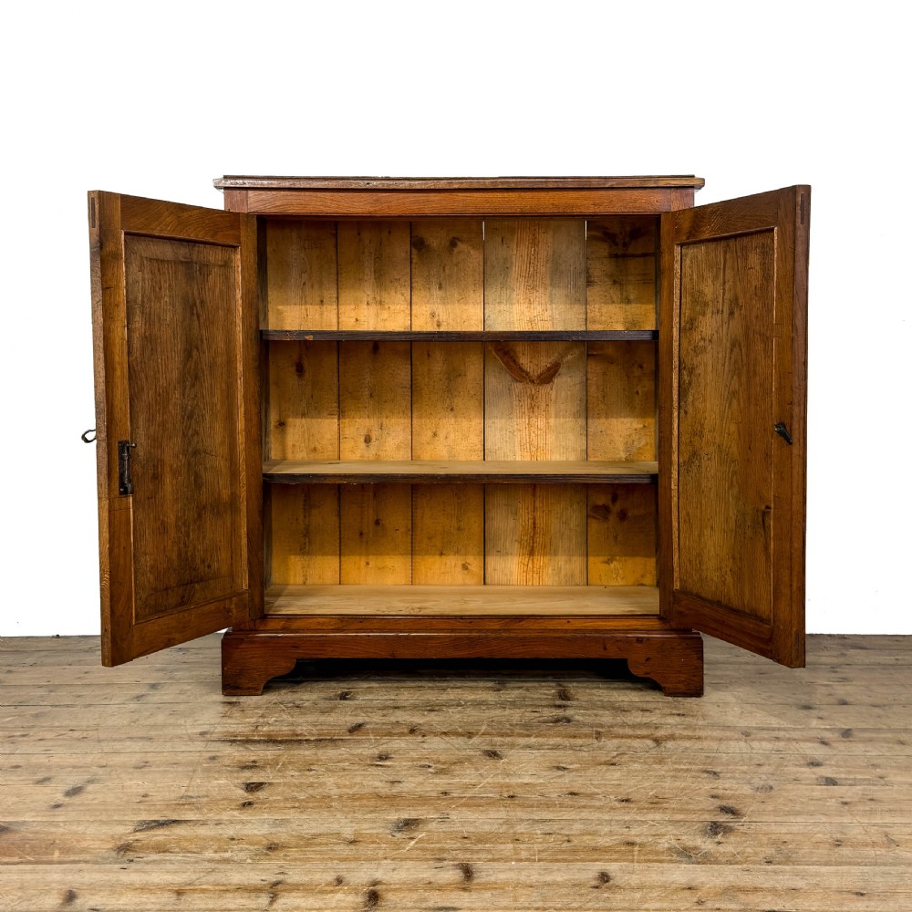An antique warm brown oak cupboard featuring two moulded panel doors, polished brass escutcheons, and a shaped plinth base. View VII
