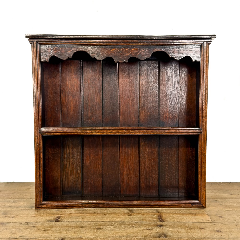 A dark brown oak two-tier wall-hanging shelf unit featuring a scalloped top frieze and vertical planked backboard.