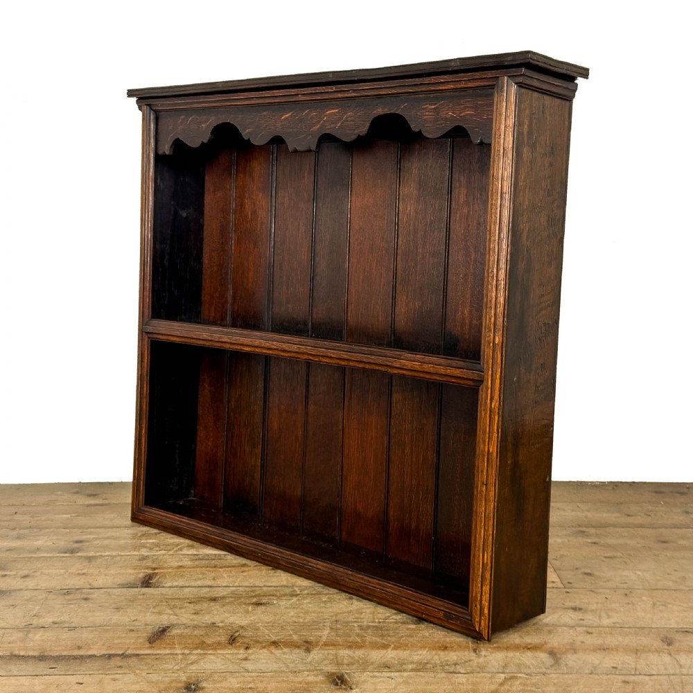 A dark brown oak two-tier wall-hanging shelf unit featuring a scalloped top frieze and vertical planked backboard. View II