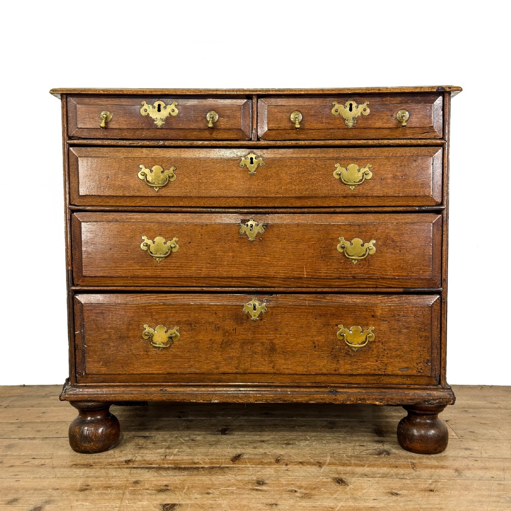 An antique 18th-century oak chest of drawers featuring a two short over three long drawer arrangement, brass handles, and turned bun feet.