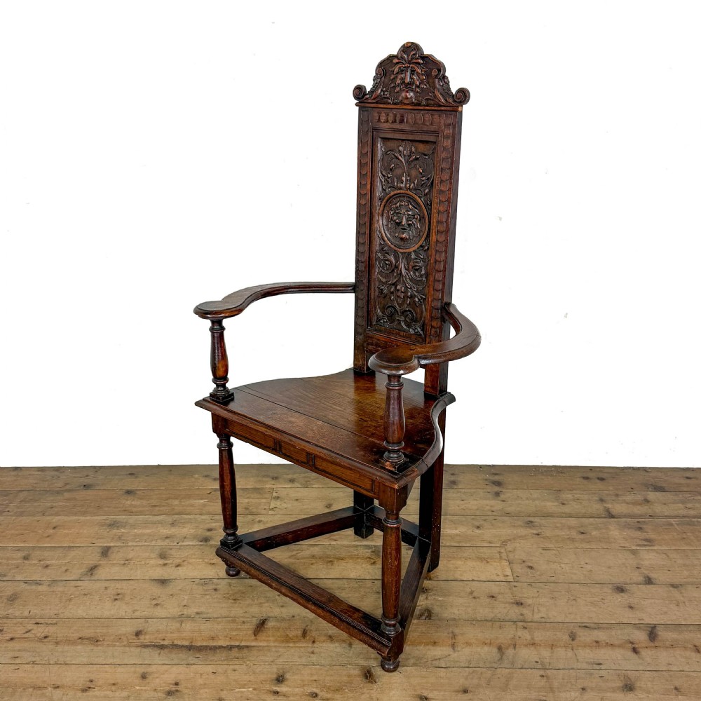 A dark carved oak high-back elbow chair featuring relief face carvings, curved armrests, turned legs, and a solid timber seat.