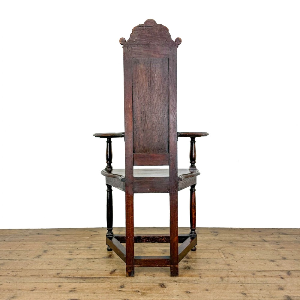 A dark carved oak high-back elbow chair featuring relief face carvings, curved armrests, turned legs, and a solid timber seat. View II