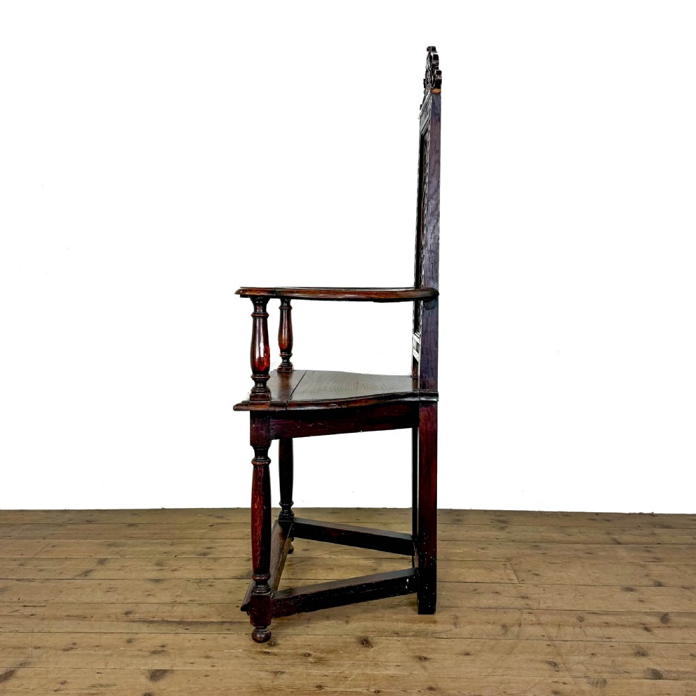 A dark carved oak high-back elbow chair featuring relief face carvings, curved armrests, turned legs, and a solid timber seat. View III