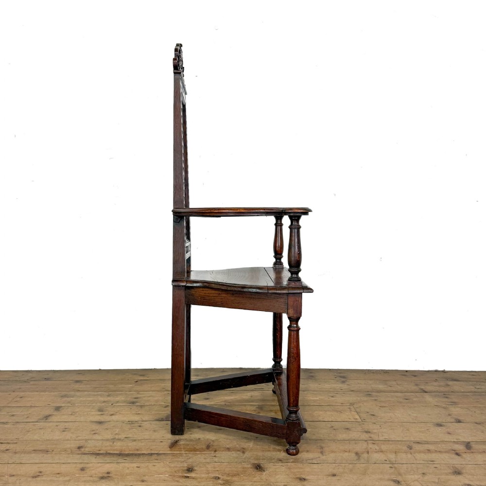 A dark carved oak high-back elbow chair featuring relief face carvings, curved armrests, turned legs, and a solid timber seat. View IV