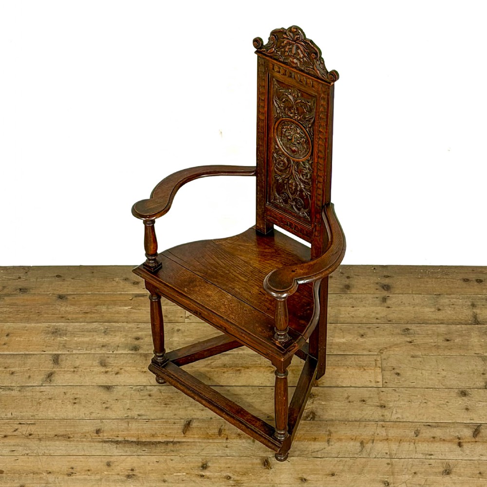 A dark carved oak high-back elbow chair featuring relief face carvings, curved armrests, turned legs, and a solid timber seat. View V