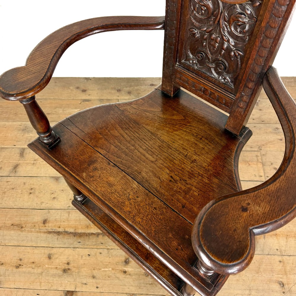 A dark carved oak high-back elbow chair featuring relief face carvings, curved armrests, turned legs, and a solid timber seat. View VI