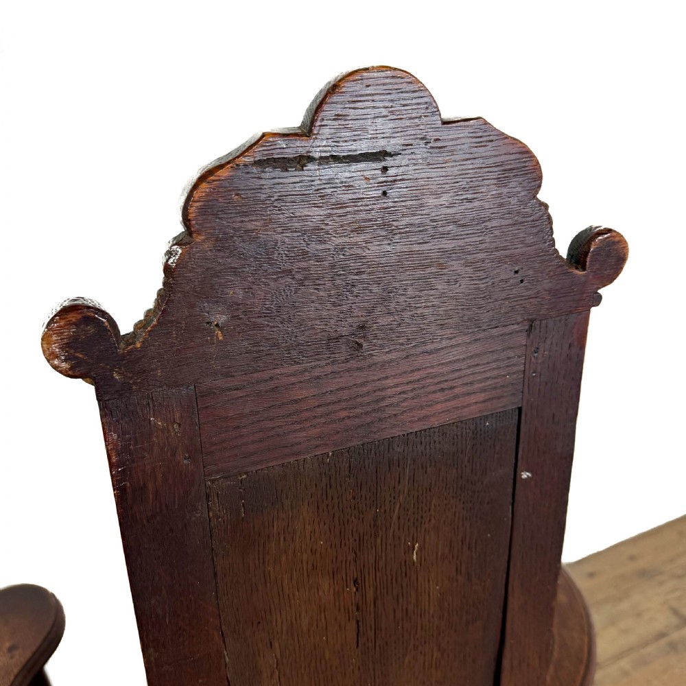 A dark carved oak high-back elbow chair featuring relief face carvings, curved armrests, turned legs, and a solid timber seat. View VII