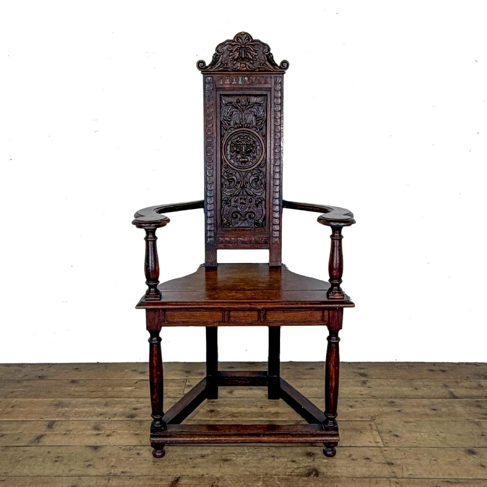A dark carved oak high-back elbow chair featuring relief face carvings, curved armrests, turned legs, and a solid timber seat. View VIII