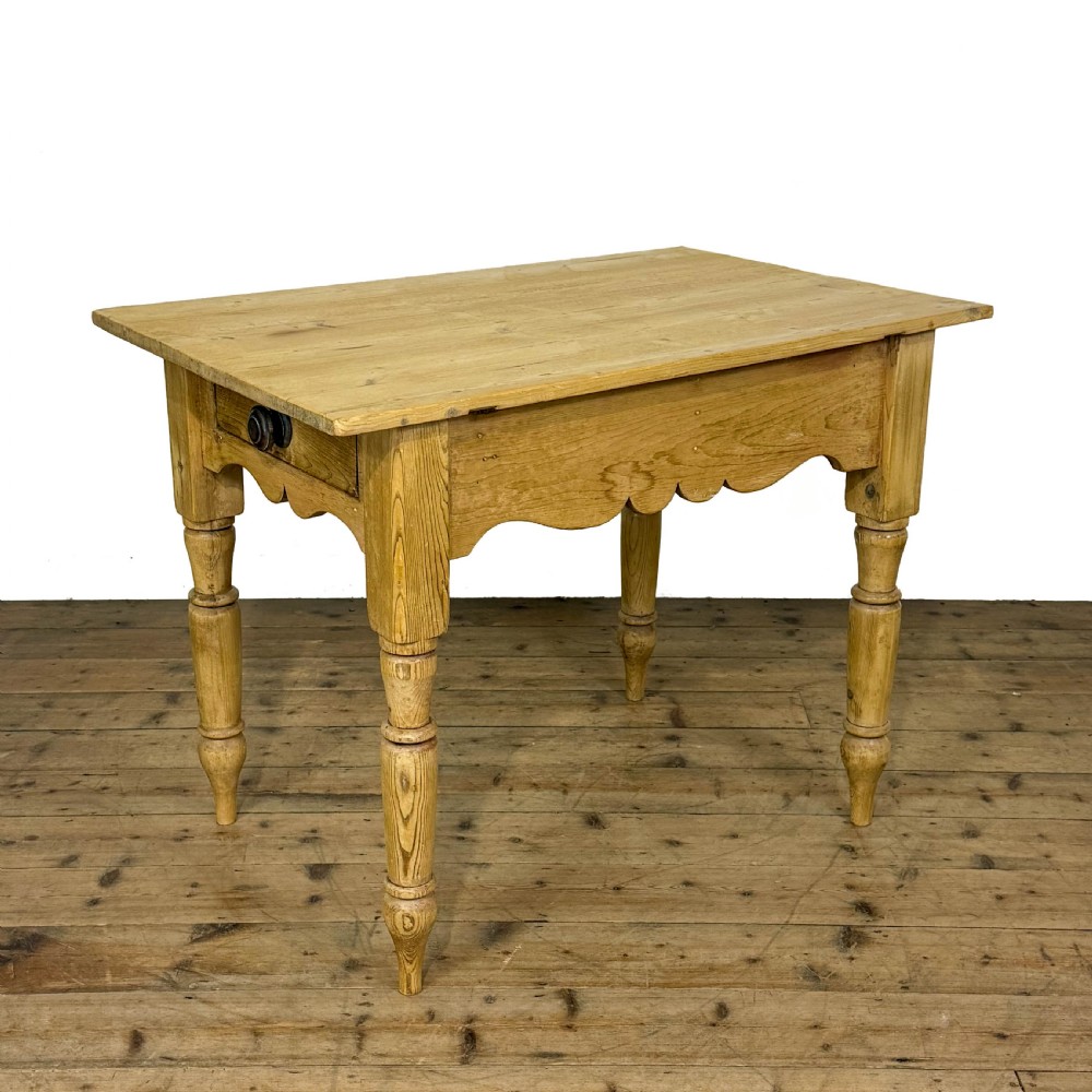 An antique honey pine kitchen table featuring hand-turned legs, a scalloped apron, and a single end drawer with a dark turned wooden knob.