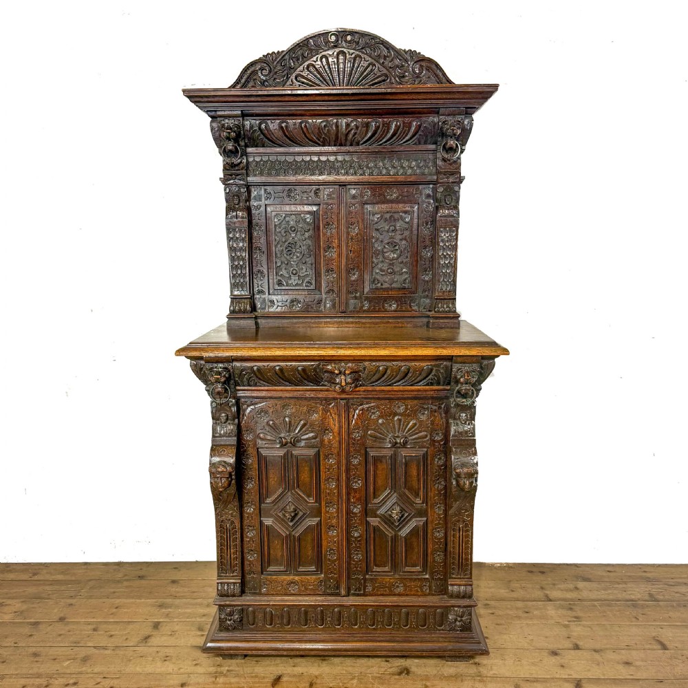 A two-tier dark oak cupboard featuring relief carvings, lion masks, geometric panelled doors, and a decorative carved pediment.