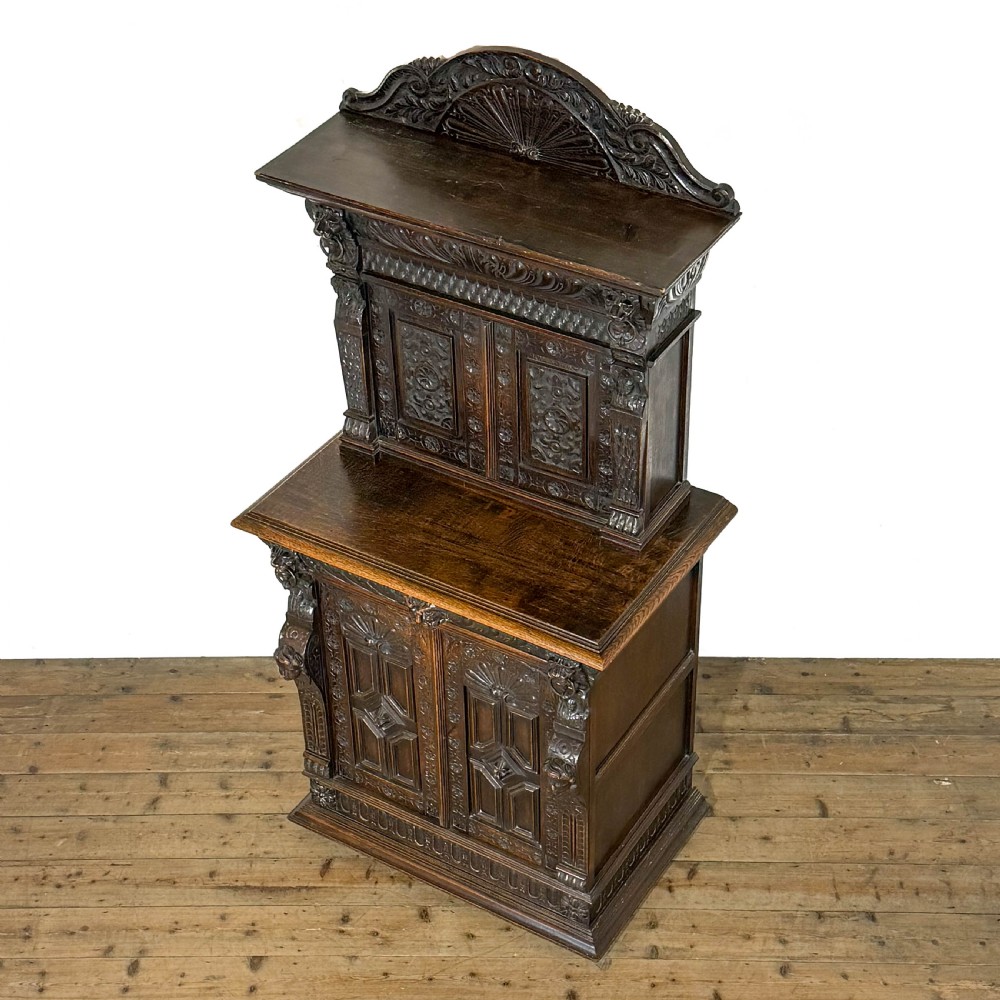 A two-tier dark oak cupboard featuring relief carvings, lion masks, geometric panelled doors, and a decorative carved pediment. View II