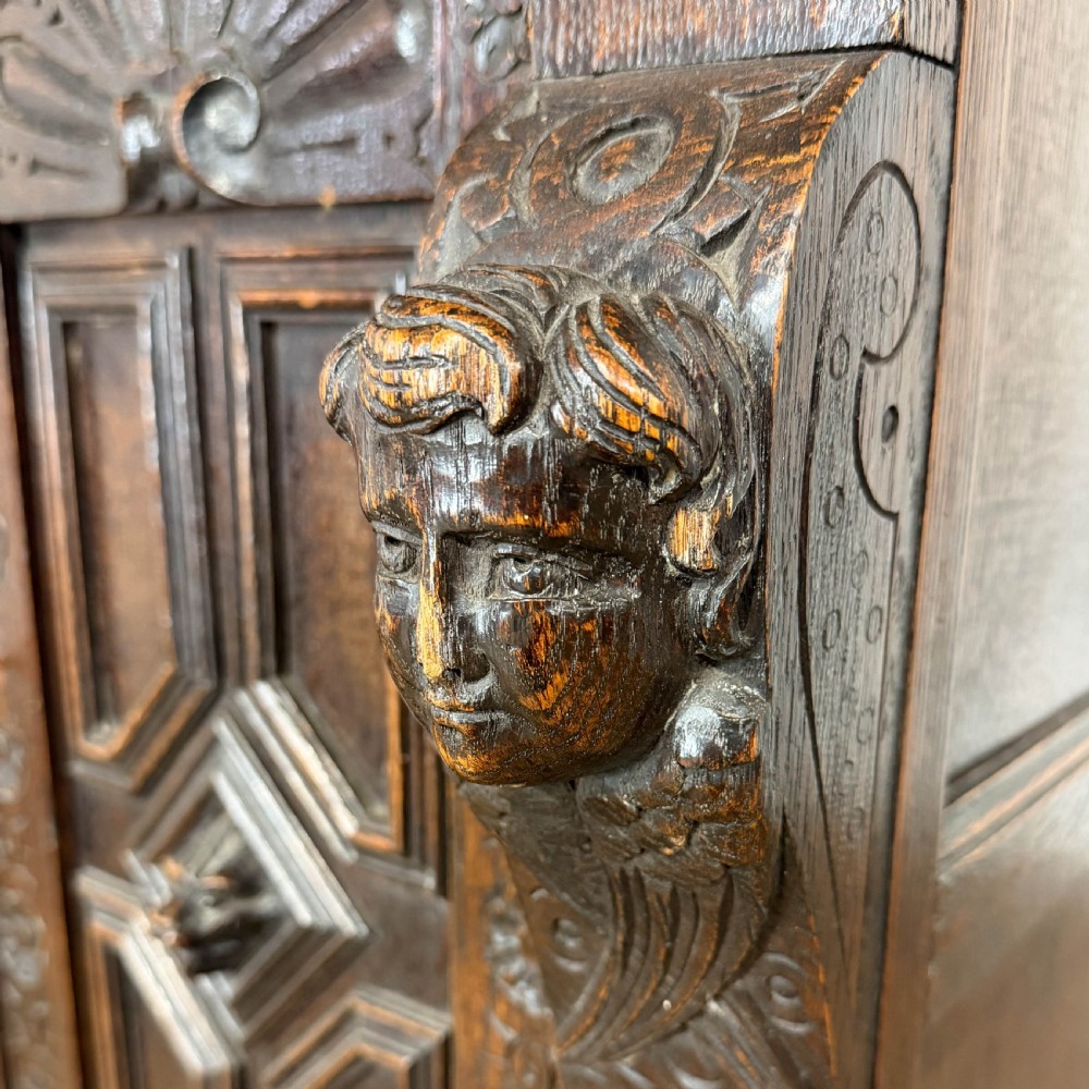 A two-tier dark oak cupboard featuring relief carvings, lion masks, geometric panelled doors, and a decorative carved pediment. View III