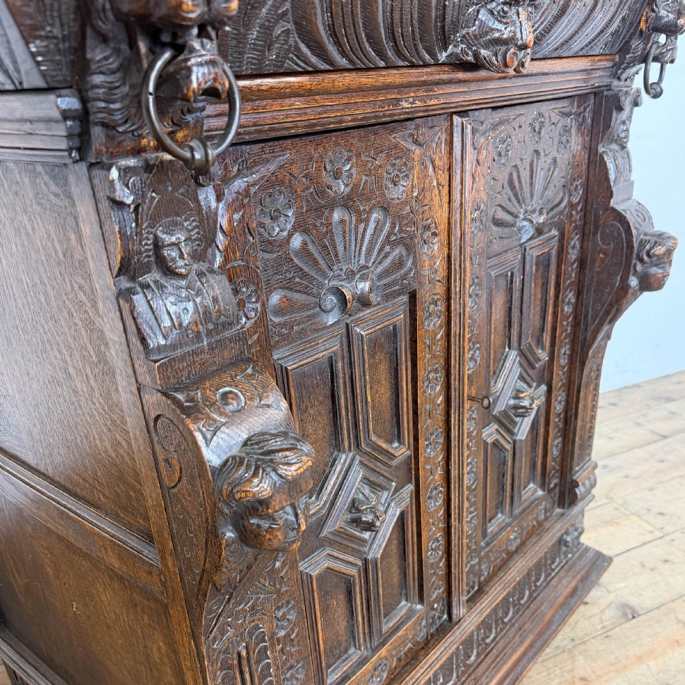 A two-tier dark oak cupboard featuring relief carvings, lion masks, geometric panelled doors, and a decorative carved pediment. View IV