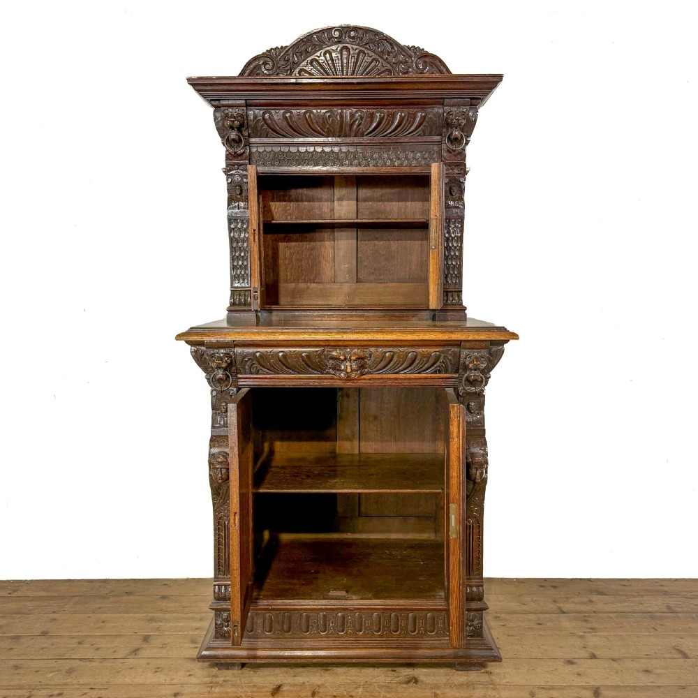 A two-tier dark oak cupboard featuring relief carvings, lion masks, geometric panelled doors, and a decorative carved pediment. View VI