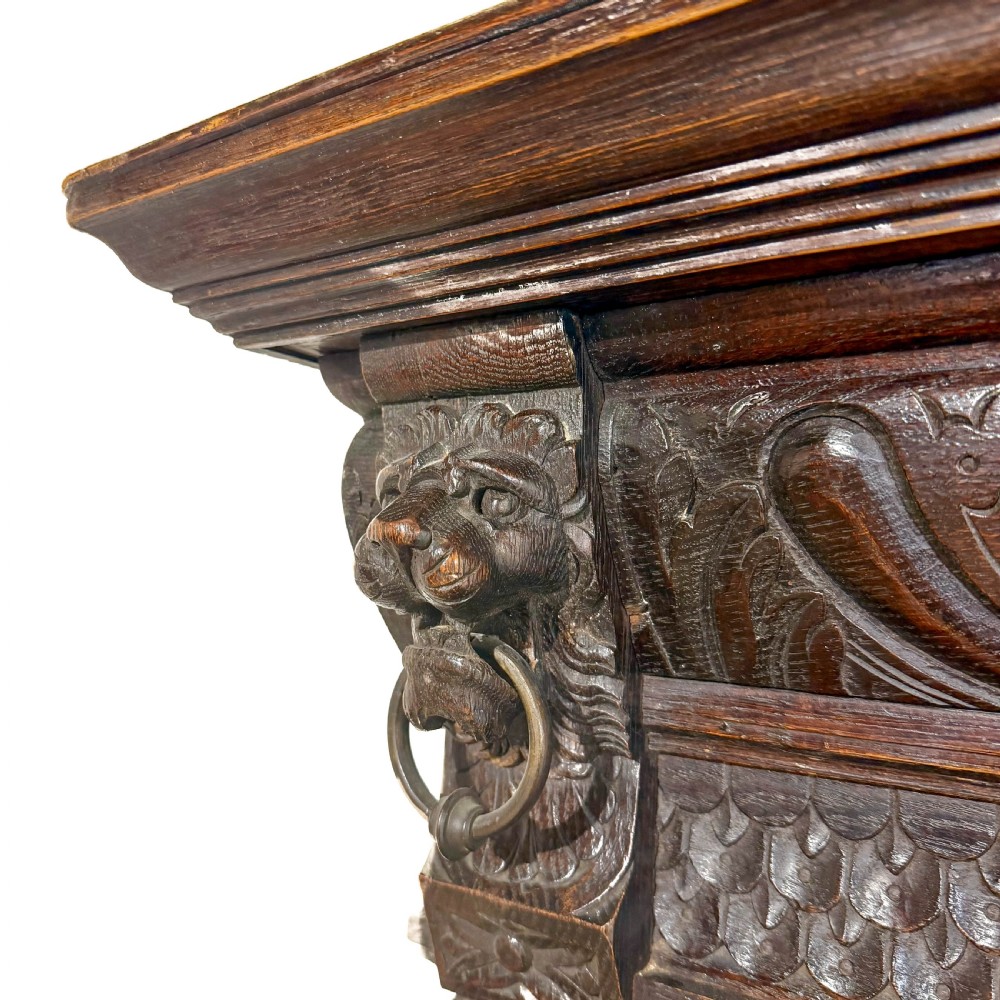 A two-tier dark oak cupboard featuring relief carvings, lion masks, geometric panelled doors, and a decorative carved pediment. View VII