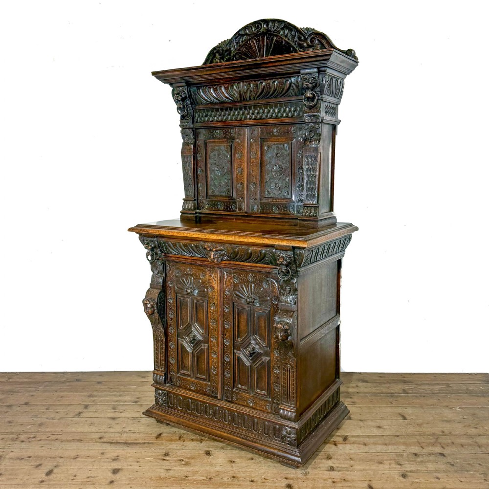 A two-tier dark oak cupboard featuring relief carvings, lion masks, geometric panelled doors, and a decorative carved pediment. View VIII