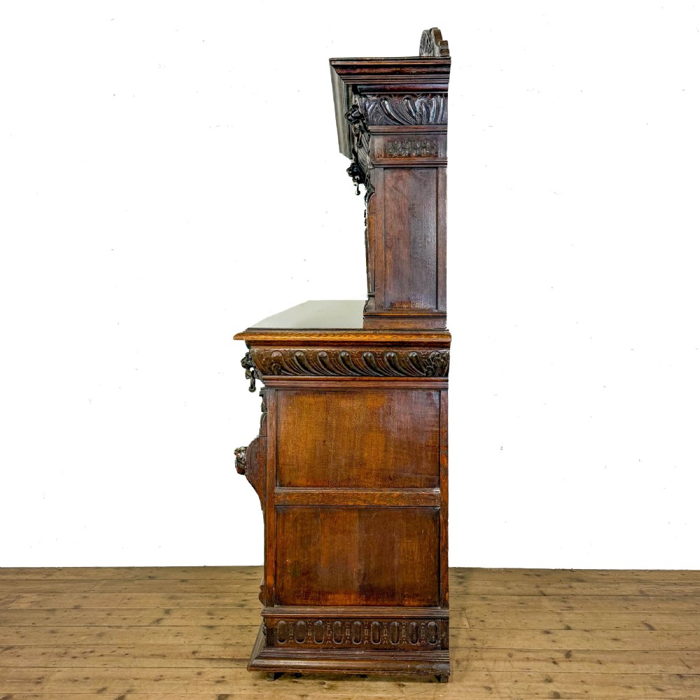A two-tier dark oak cupboard featuring relief carvings, lion masks, geometric panelled doors, and a decorative carved pediment. View IX
